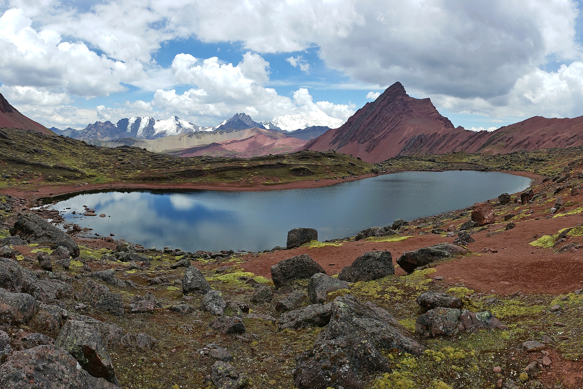 A beautiful alpine lake in the Ausangate region of Peru, surrounded by bright green lichen, red rocky peaks, and jagged, glaciated peaks in the distance