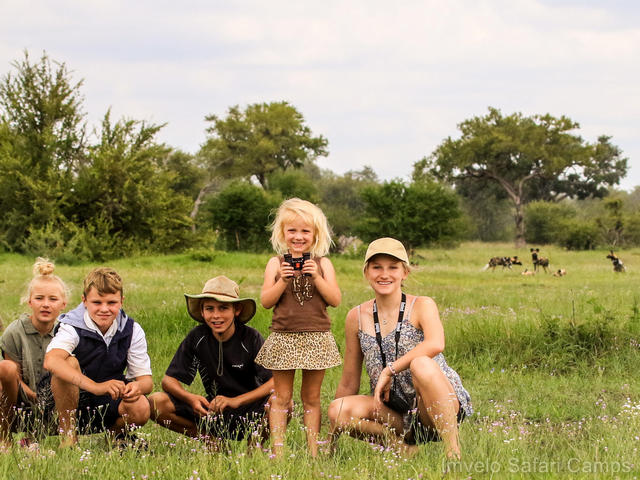 A family with young children smiling in the grass, with African wild dogs in the distance behind them, on a walking safari in Hwange National Park