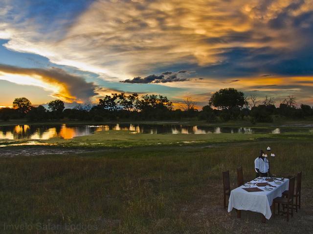 An outdoor dining table set with white linens under a colorful sunset at a luxury safari camp in Hwange National Park Zimbabwe