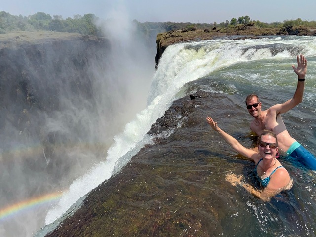 Two smiling guests wave their arms from the dropoff at the very edge of Victoria Falls at the Devil's Pool