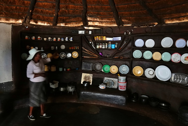 inside a thatch-roofed hut in Ngame village, with wooden shelves displaying cooking pots, plates, and other kitchenwares
