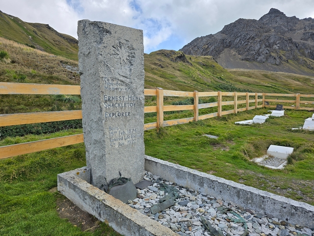 the gray, rectangular grave of Ernest Shackleton, with the mountains of Gritviken South Georgia Island visible in the distance